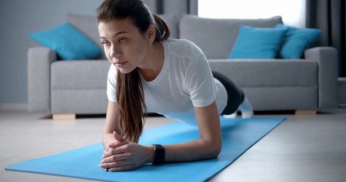 Concentrated beautiful woman in sport clothing doing plank exercise on blue yoga mat in living room. Slim fitness girl with dark hair having abs workout at home.