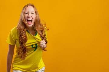 Brazil supporter. Brazilian redhead woman fan celebrating on soccer, football match on yellow background. Brazil colors.