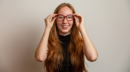 Beautiful young redhead girl in eyeglasses is looking at camera and smiling, on gray background.