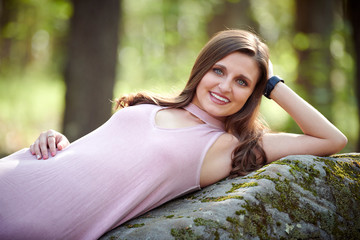 Gorgeous young Caucasian woman in pink cocktail dress stands in summer forest setting - laying on boulder