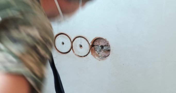 Male builder works at construction site, repairing room, making holes in brick wall with drill, place for electrical outlets, brick fragments, pulling out pieces with his hands, drill, perforator