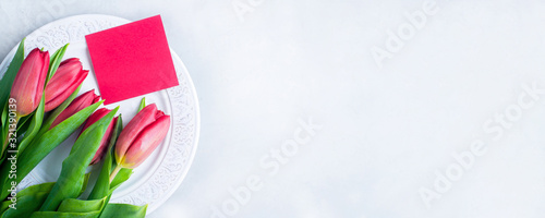 Festive Table Setting with pink tulips and red card on white plate. Concept Spring, Easter or Mother's Day. Flat lay, top view, copy space. Banner image for website.