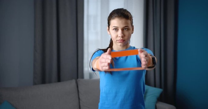 Strong young woman with concentrated facial expression in sports wear doing exercises for hands by rubber fitness band at home. Charming lady enjoying workout indoors