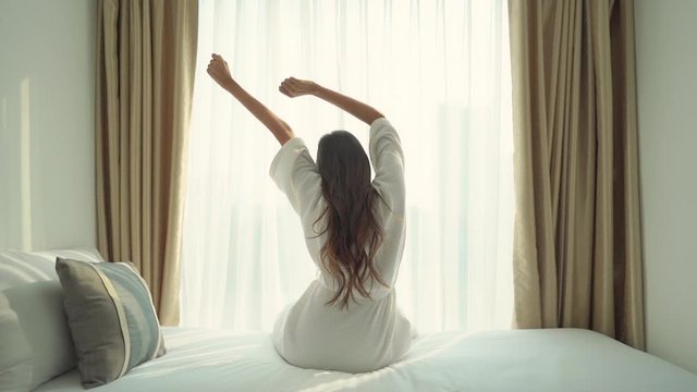 Beautiful brunette woman sitting on bed stretching arms on bathrobe, bright window background