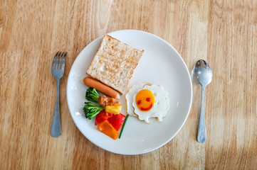 A white dish of kid's breakfast decorated with sheep smiling omelette, grilled bread, sausage and vegetable on wooden table with fork and spoon, top view image