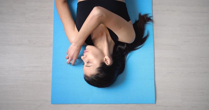 Fitness young woman with dark hair and bare torso taking wireless earphone from blue yoga mat for listening favorite music during training. Concept of sporty lifestyle. Top view.