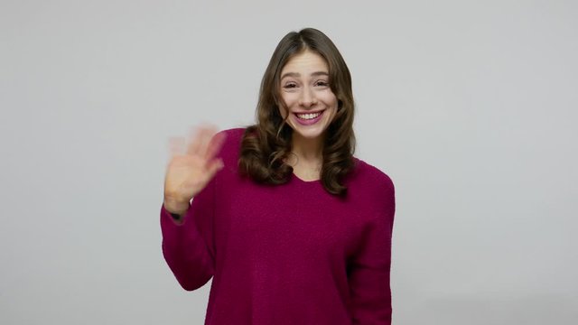 Hello! Charming nice-looking brunette woman in pullover waving hand gesturing hi to camera, welcoming with friendly engaging smile, expressing joy. indoor studio shot isolated on gray background