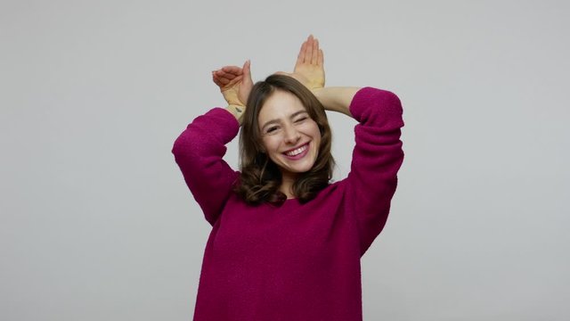 I'm rabbit! Amusing childish nice-looking woman in pullover showing bunny gesture and making faces, having fun with cute playful comical expression. indoor studio shot isolated on gray background