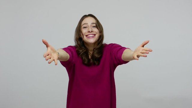 Come into my arms! Charming friendly hospitable brunette woman in pullover holding hands wide open to hug you, welcoming with kind smile, going to embrace. studio shot isolated on gray background
