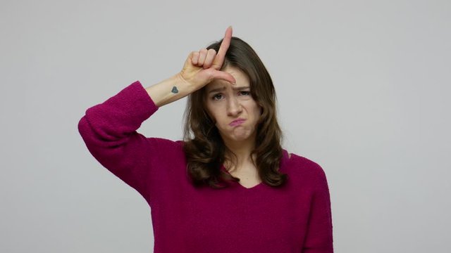 Unhappy brunette woman in pullover showing L sign with fingers, gesturing I'm looser, feeling frustrated depressed about defeat, unsuccess, lost job. indoor studio shot isolated on gray background