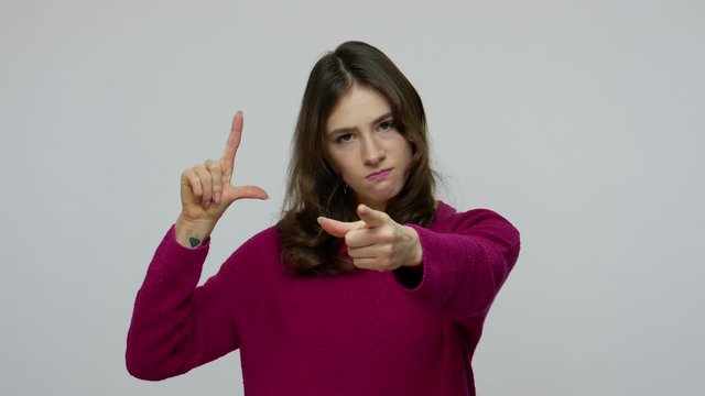 Angry brunette woman showing L sign with fingers, gesturing You are loser and pointing to camera, blaming for defeat unsuccess, dismissal from work. indoor studio shot isolated on gray background