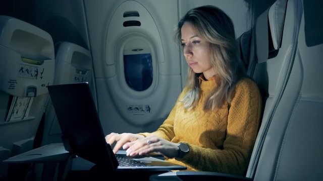 Female freelancer working on laptop sitting In airplane. A lady is working on a laptop in semi-darkness while being on a plane
