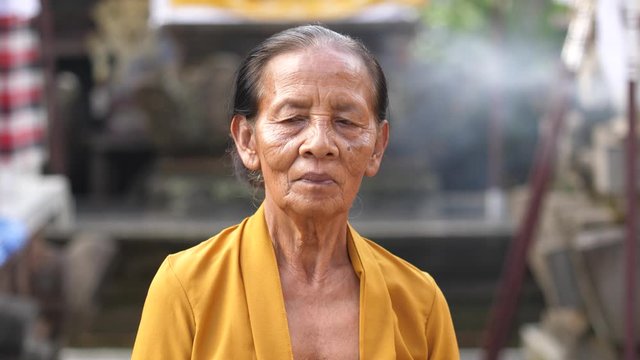 Elderly Asian woman stands in courtyard of Hindu temple in Bali with smoke from incense blowing in background. Old, weathered Balinese lady in bright yellow smiles at camera.