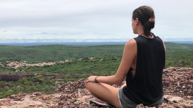 Young woman meditates on top of a rock mountain with beautiful forest and small town landscape ahead