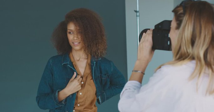 Young professional female photographer taking photos of a beautiful african american woman modeling for a luxury clothing brand, behind the scenes of a studio fashion photoshoot