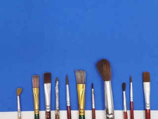 A row of eleven different painting brushes on a blue paper. Art supplies / instruments / tools. Creative process concept.