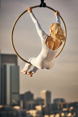 Young Woman Sitting in a Large Hoop Suspended in Air with Dramatic Cityscape in the Background in Golden Hour Sunset