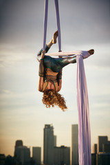 Young Woman Performing Aerial Silk Dance while Suspended in Air with Dramatic Cityscape in the Background in Golden Hour Sunset