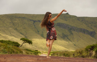Young woman in dress turning head backwards with raised hand on country road. View from behind. Good bye hand in the air