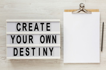 'Create your own destiny' words on a lightbox, clipboard with blank sheet of paper on a white wooden background, top view. Overhead, from above, flat lay. Copy space.