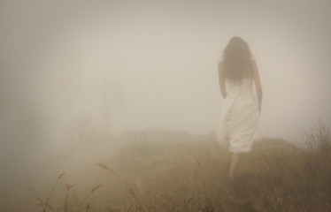 Fototapeta premium Kobieta w długiej białej sukni w mglistej mgle. Rozmyć dostrzegalny zarys sylwetki. Widok od tyłu. Tajemniczy krajobraz