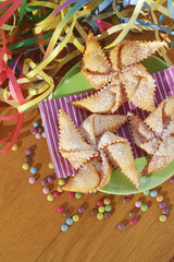 Traditional carnival pastry, chiacchere or crostoli, bugie, cenci in shape of a toy windmill on wooden background with coloful decorations