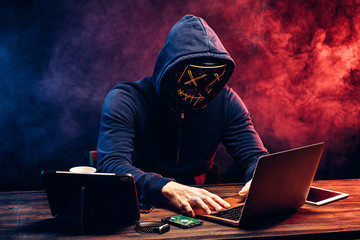 hacker man typing on laptop, hacking computer system. male in mask and pullover. unrecognizable incognito male sit in hood and try to breach the security of laptop system. neon smoke in background - obrazy, fototapety, plakaty