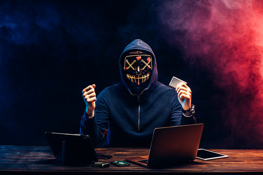 anonymous hacker in mask after successful getting access to bank card, raised hands up, hold bank card in one hand, going to withdraw money from it. isolated over black background
