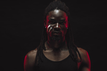 confident muscular black man isolated over black background. male in black shirt, having dreadlocks on head and strips on cheek like an indian man look at camera, red neon rays fall on the face