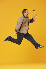 Fototapeta na wymiar Side view full length of joyful mature worker jumping high holding hammer against yellow background in studio, 90s video game style, copy space