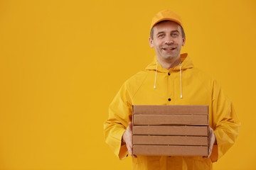 Waist up portrait of adult delivery man holding pizza boxes and smiling at camera while standing against pop yellow background, copy space