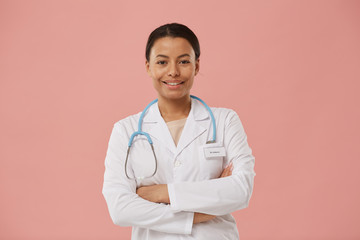Waist up portrait of beautiful female doctor smiling at camera while standing with arms crossed against pale pink background, copy space