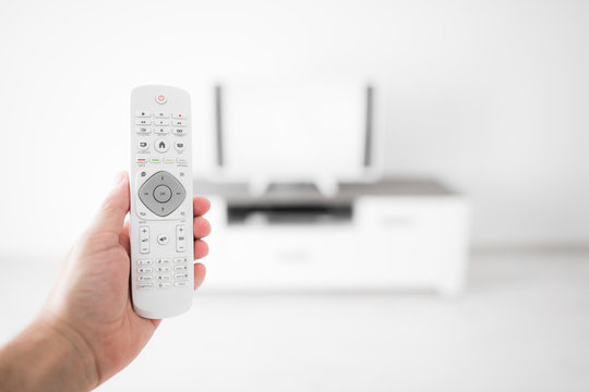 Hand holding TV remote controller with a modern television set.