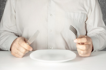 A man without a face sits at a table with a knife and a fork in his hands in front of an empty white plate.