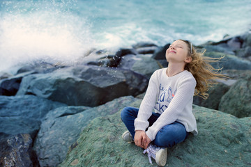 a 9-year-old girl with long white hair sits on the seashore on the rocks, waves crash on the rocks