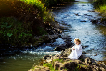 Horizontal portrait of 5 years old girl in white traditional embroidery ukrainian dress and chamomiles wreath on rocks in river in Carpathian mountains. Travel tourism destination in Ukraine.