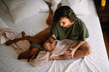 Girl with dog in bed. Woman drinking coffee and using phone. 