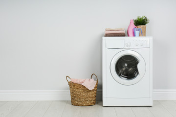 Modern washing machine with stack of towels, detergents and laundry basket near white wall. Space...