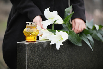 Woman holding white lilies near black granite tombstone with candle outdoors, closeup. Funeral...