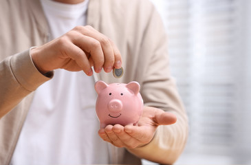 Man putting coin into piggy bank on light background, closeup