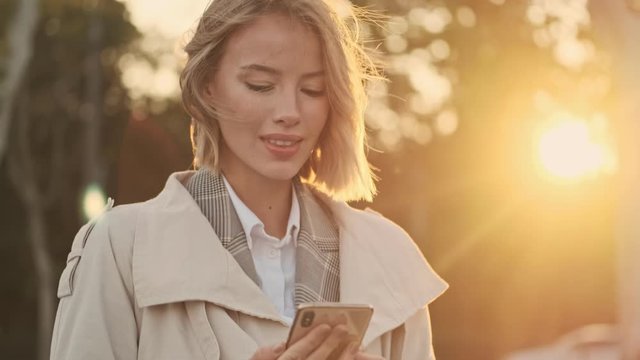 Pleased blonde business woman in coat using smartphone on parking near airport outdoors
