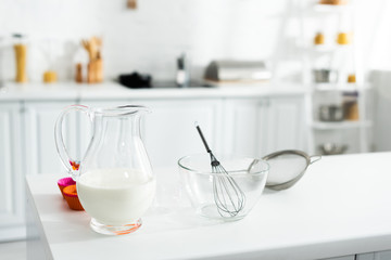 jug with milk, bowl, balloon whisk, sieve on table in kitchen