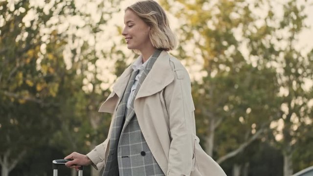 Side view of Cheerful blonde business woman in coat walking with luggage on airport parking outdoors