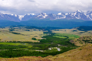 Fototapeta premium Panoramic view of mountain range under cloudy sky. Plateau with green trees and small rivers against snow-capped peaks.
