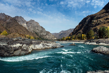 Mighty mountain river flows along stony banks among rocky mountains against a clear blue sky. Turquoise water of stormy river and huge stones.