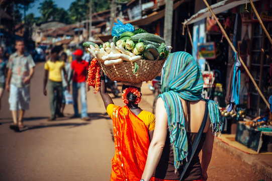 An Indian woman in red Sari carries a large basket of vegetables on her head. Tourists and locals in India. A crowded street in Gokarna, Karnataka.