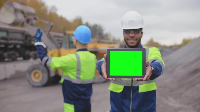 Industrial worker looking at camera and showing digital tablet screen on construction site