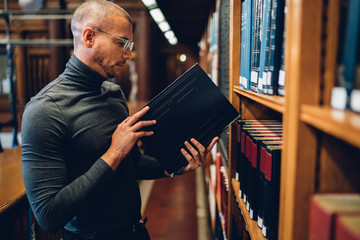Serious male student standing near bookshelves checking literature for researching information, concentrated smart historian holding book in public library working with scientist publication