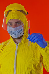 Man in yellow chemical protection suit and face protective mask coronavirus. Without glasses. Protective helmet. Hands in blue gloves. Syringe with red liquid.
