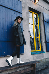 Fototapeta na wymiar Long haired blond woman wearing a black fedora hat, a black coat with woollen lapels, and white boots, standing on a wooden bench behind blue window shutters